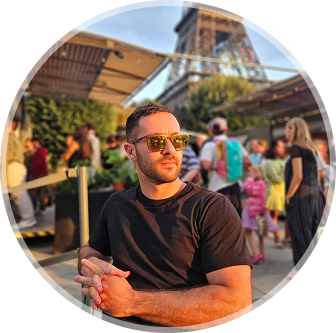 Imagem de João Damiani sério de camisa preta em Paris sentado abaixo da Torre Eiffel